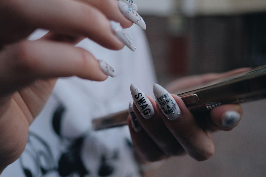 Unhas acrílicas femininas com strass e texto.